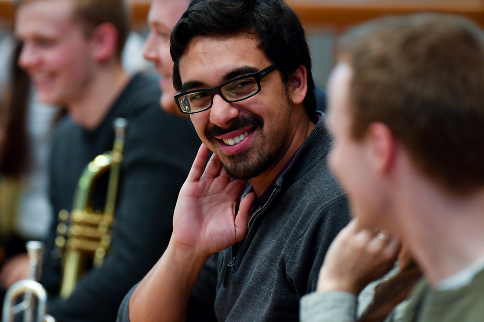 A male student, with dark hair and glasses, smiling at another student, while they are waiting further instruction in a orchestra rehearsal.